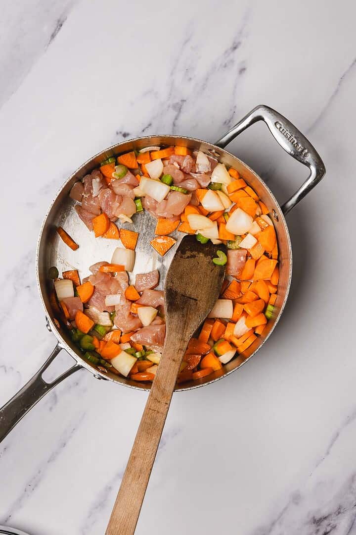 A skillet with chopped chicken, carrots, onions, and celery being stirred with a wooden spoon on a marble surface—perfect for easy chicken recipes like classic chicken and dumplings.