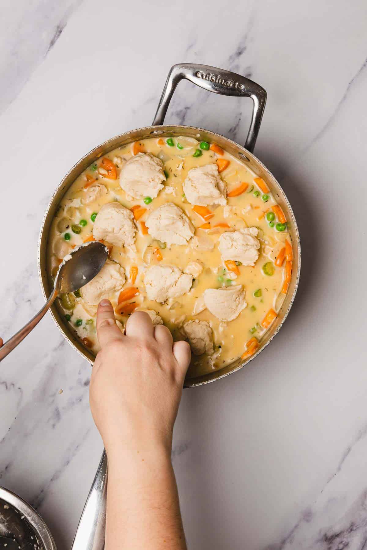 A hand placing biscuit dough onto a pot of creamy vegetable and chicken stew on a marble countertop, creating an easy Chicken and Dumplings recipe.