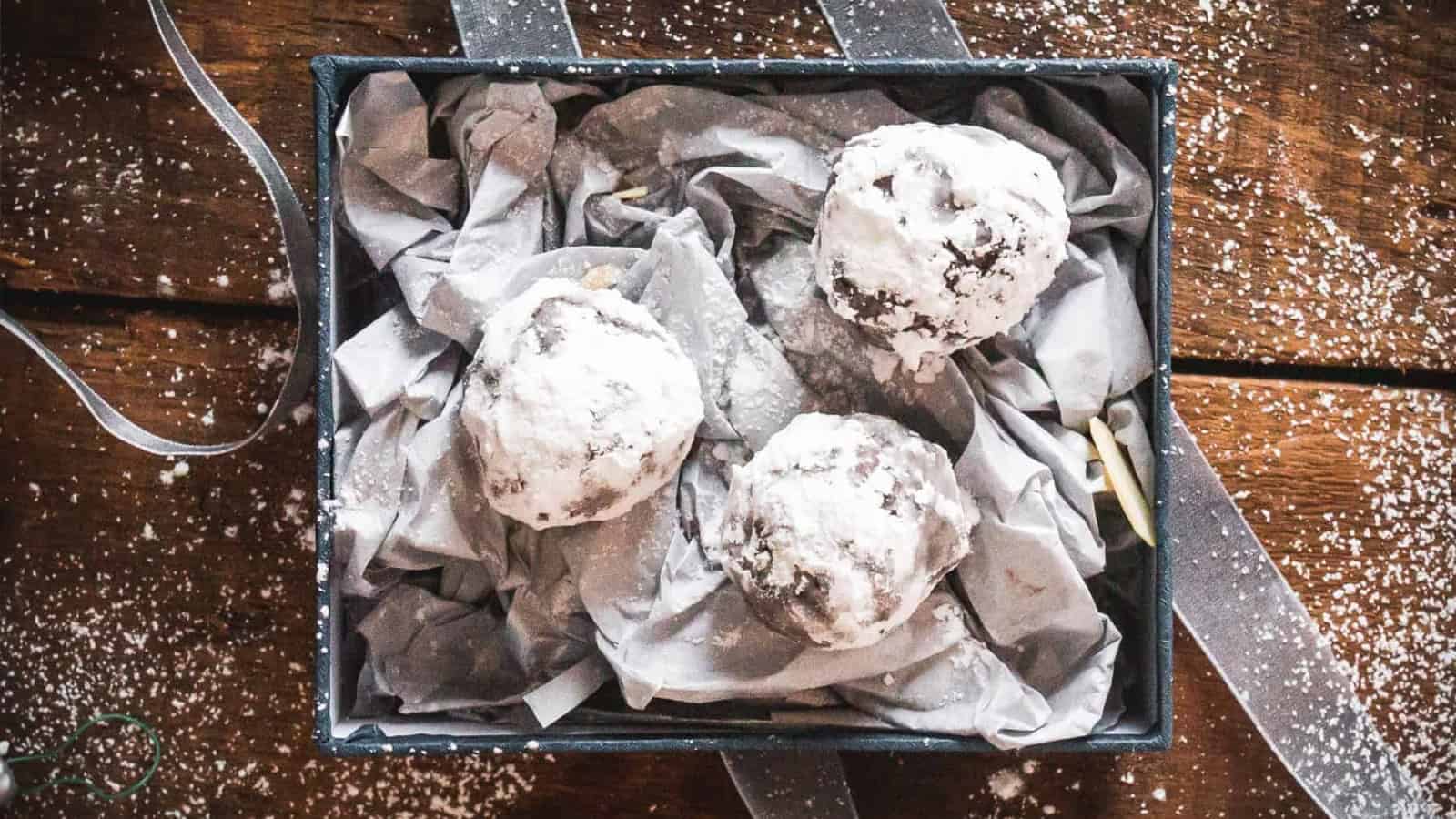 Three powdered sugar-coated cookies in a box lined with crumpled gray tissue paper on a wooden surface.