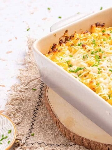 A baked casserole topped with melted cheese and chopped chives in a white dish on a wooden board.