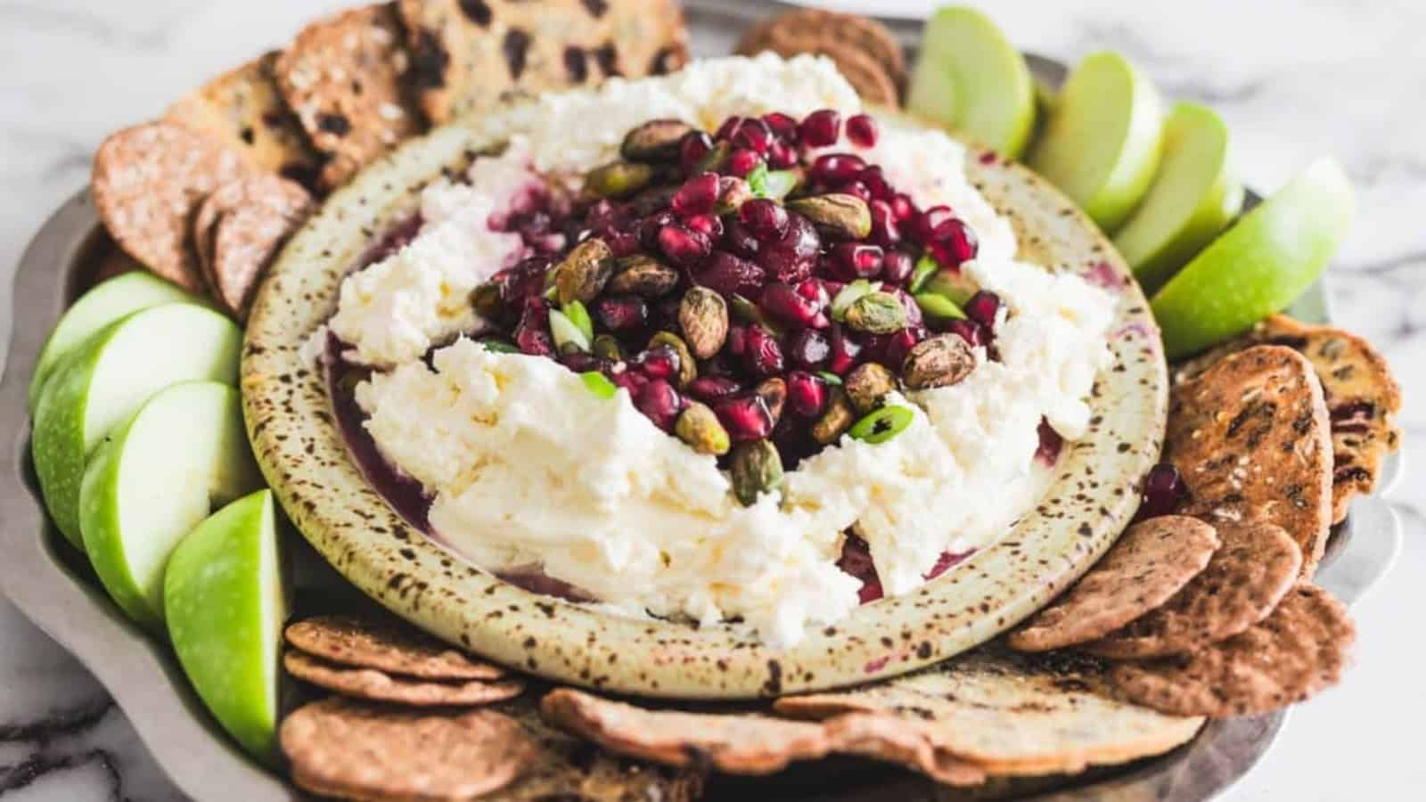 A platter with whipped cheese topped with pomegranate seeds, pistachios, apple slices, and crackers.