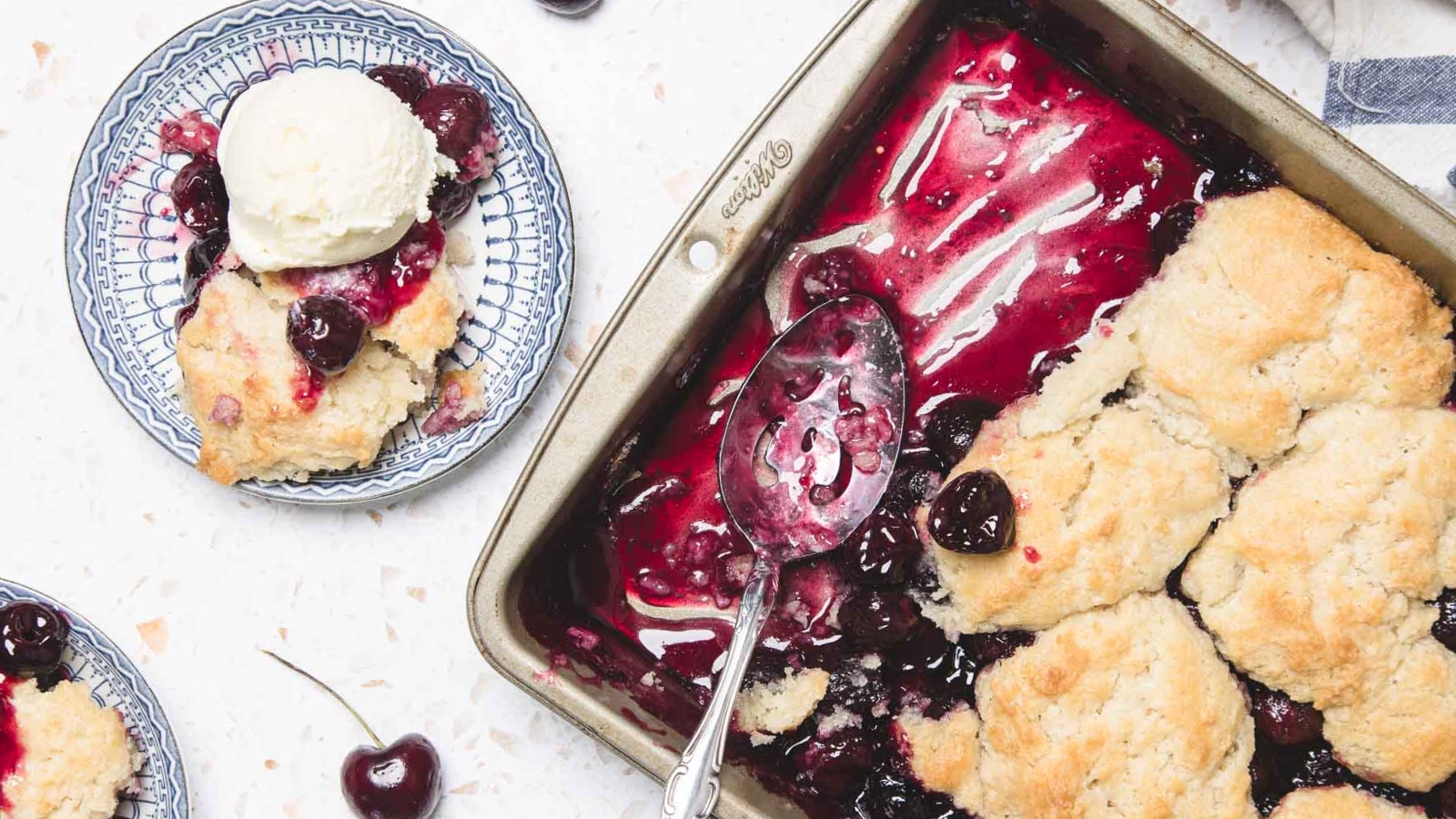 A cherry cobbler in a baking dish with a serving on a plate topped with a scoop of vanilla ice cream.