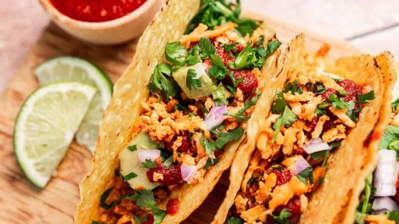Three loaded tacos with crispy shells, fresh cilantro, onions, and shredded filling, with lime wedges and salsa.