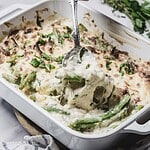 A spoon scoops cheesy green bean casserole, made with fresh green beans and topped with melted cheese and herbs, from a white baking dish.