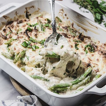 A spoon scoops cheesy green bean casserole, made with fresh green beans and topped with melted cheese and herbs, from a white baking dish.