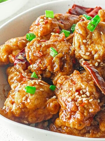 A bowl of sesame chicken garnished with chopped green onions and sesame seeds.