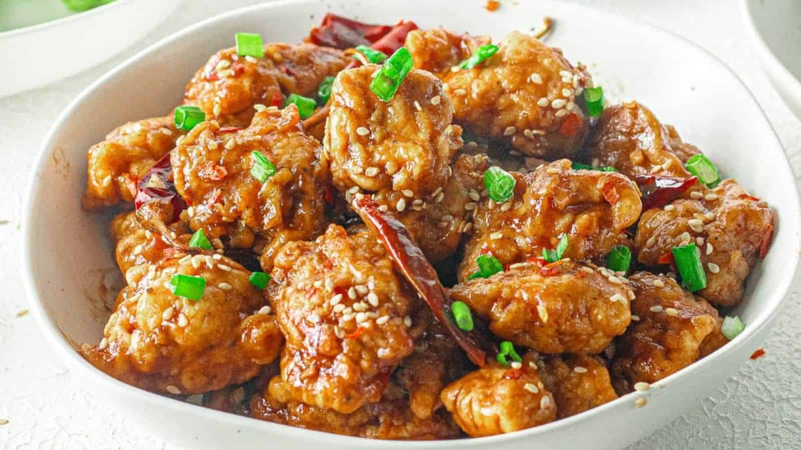 A bowl of sesame chicken garnished with chopped green onions and sesame seeds.