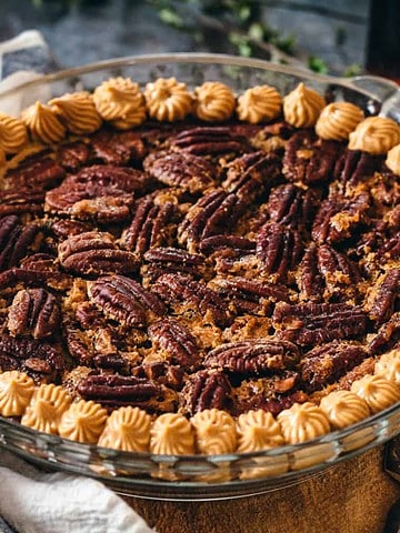 A pecan pie in a glass dish, topped with piped cream, sits on a cloth with a pie server nearby.