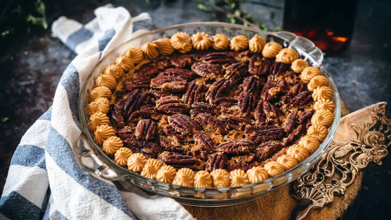 A pecan pie in a glass dish, topped with piped cream, sits on a cloth with a pie server nearby.