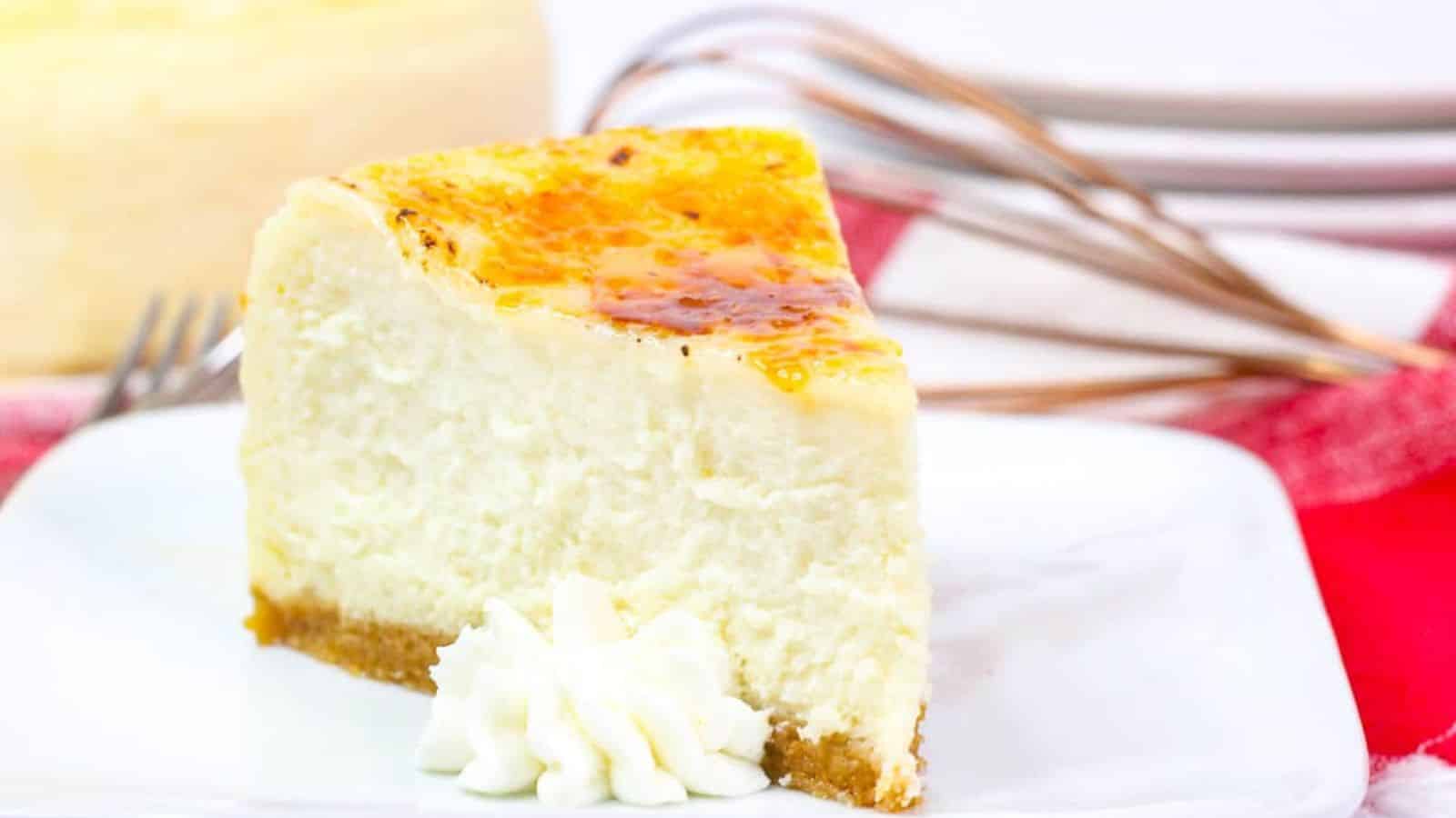 A slice of cheesecake with a caramelized top and a whipped cream dollop on a white plate, placed on a red and white checkered cloth. Plates and forks are visible in the background.