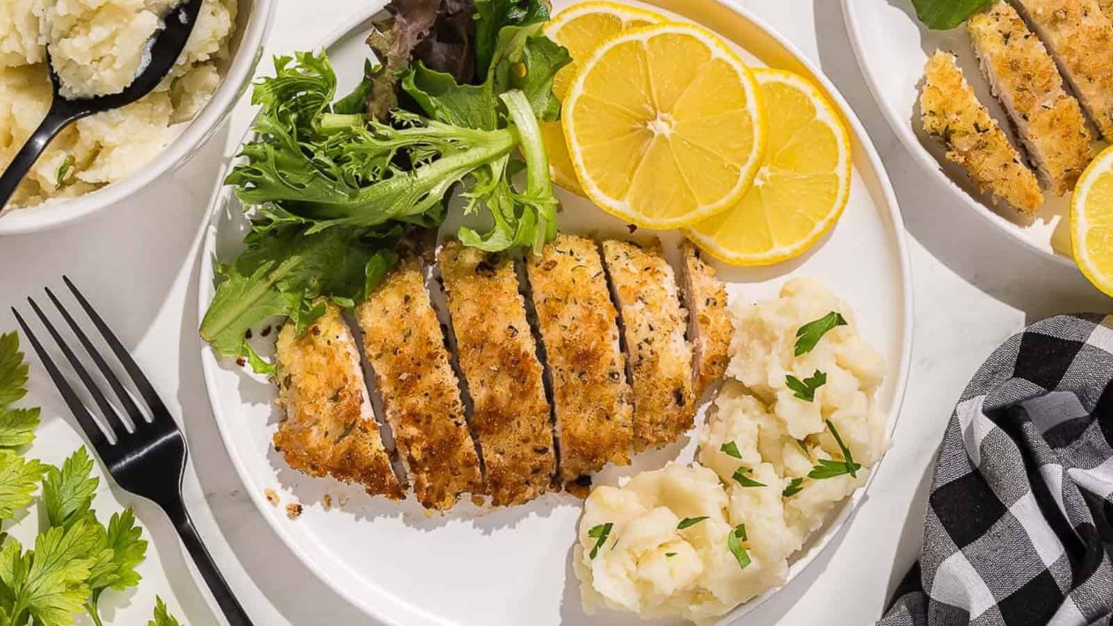 Breaded italian chicken cutlets on a black plate with sliced lemon.
