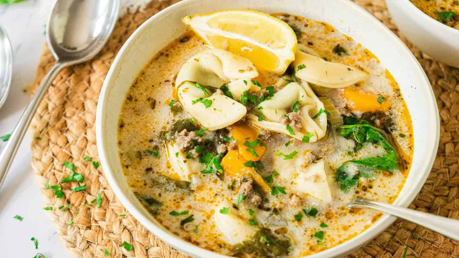 A bowl of creamy tortellini soup with spinach, lemon wedges, and herbs, served with a spoon.