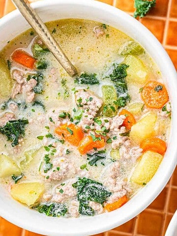 Bowl of creamy soup with sausage, carrots, potatoes, and kale, with a spoon on an orange tiled surface.