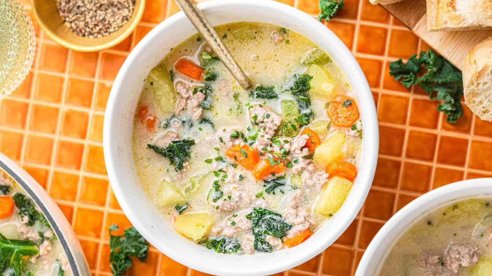 Bowl of creamy soup with sausage, carrots, potatoes, and kale, with a spoon on an orange tiled surface.