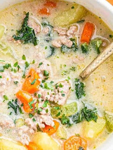 Bowl of creamy soup with ground meat, carrots, potatoes, kale, and herbs on an orange tiled surface.