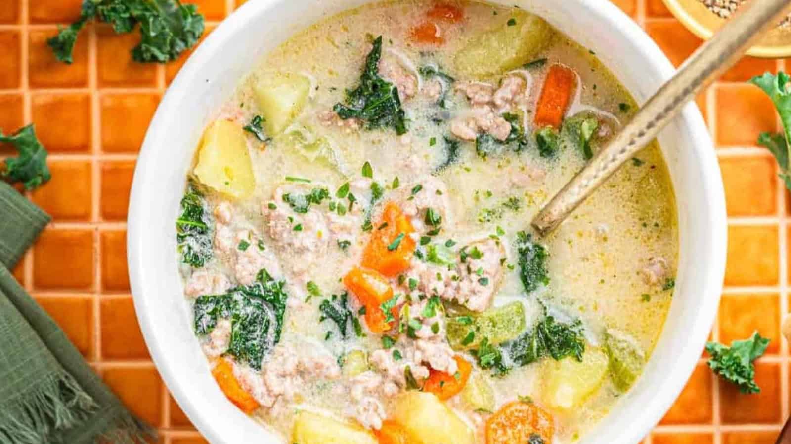 A bowl of creamy soup with sausage, kale, carrots, and potatoes, with a spoon inside, on an orange tiled surface.