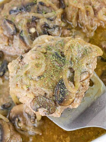 A serving spoon lifts a hamburger steak with mushrooms and onions from a baking dish with gravy.