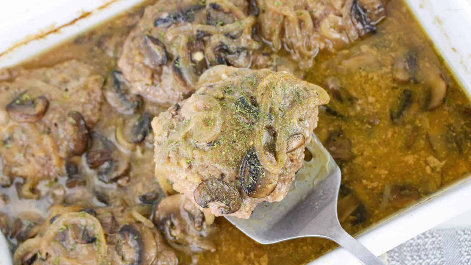 A serving spoon lifting Salisbury steak with mushrooms and onions from a baking dish with sauce.