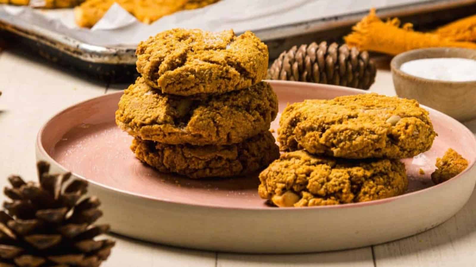 Three stacked pumpkin cookies and one beside them on a pink plate, with pine cones and a baking tray in the background.