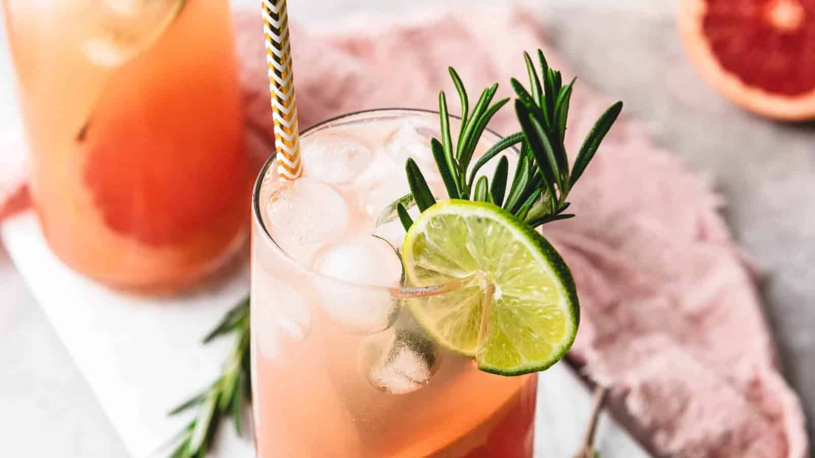 Two grapefruit cocktails with ice, lime slices, and rosemary garnish on a marble board, with a grapefruit in back.