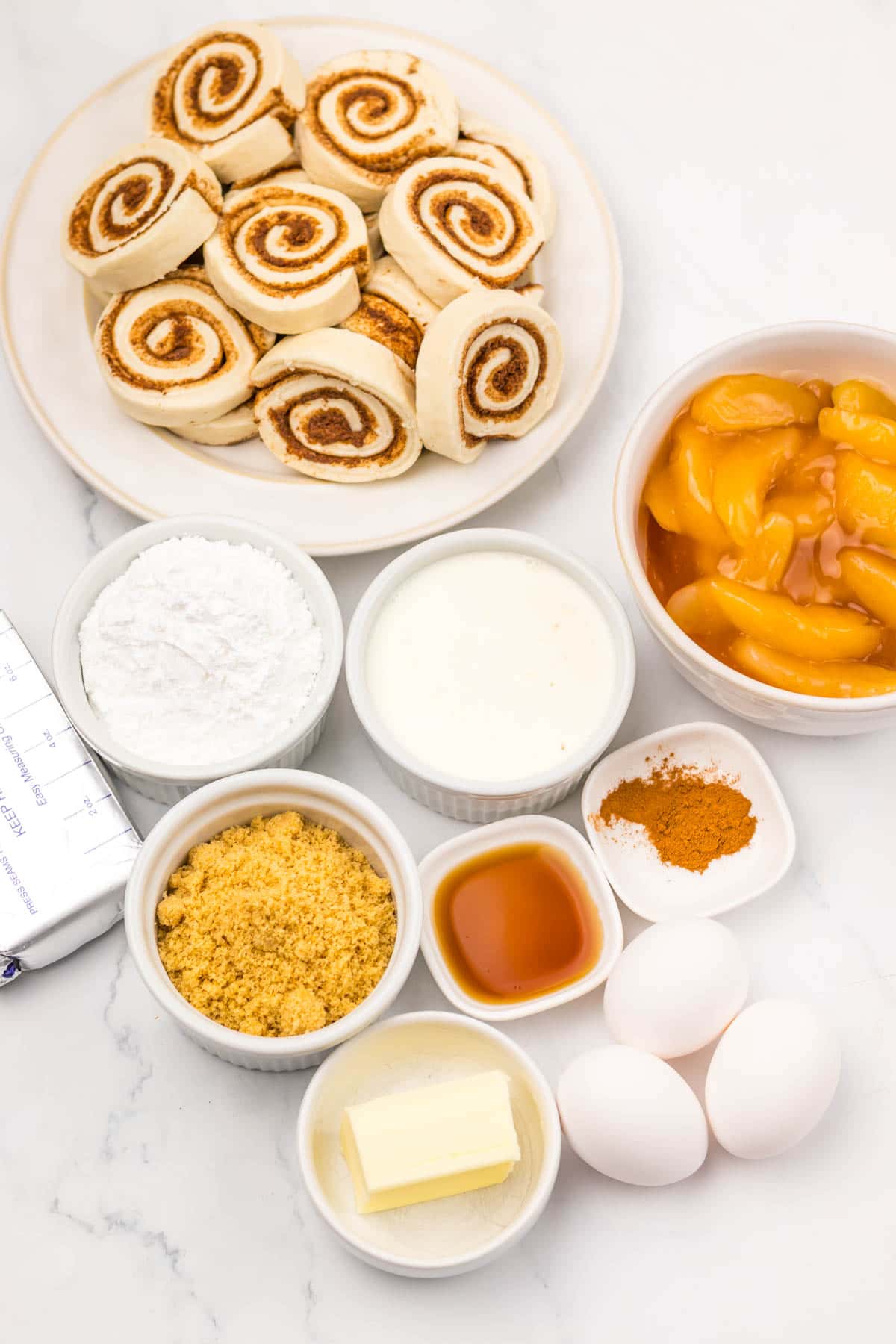 Sliced cinnamon rolls, canned peaches, eggs, butter, sugar, cream, flour, cinnamon, and vanilla on a white surface&mdash;perfect for making a slow cooker apple pie cinnamon roll casserole.