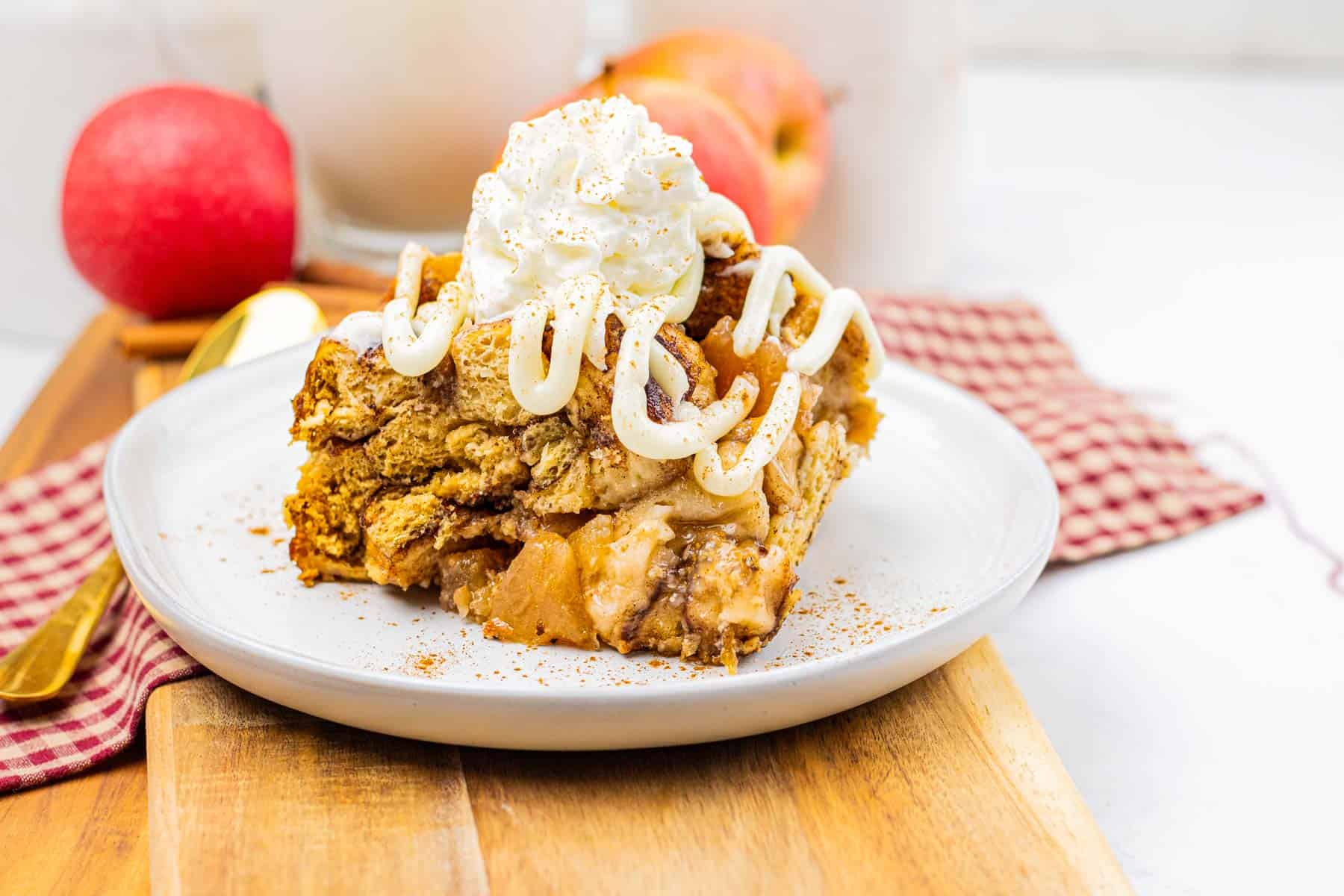 A slice of slow cooker apple pie cinnamon roll casserole topped with whipped cream and icing on a plate, with apples in the background.