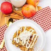 A plate of slow cooker apple pie cinnamon roll casserole with icing and whipped cream, next to apples, cinnamon sticks, and a glass of milk.