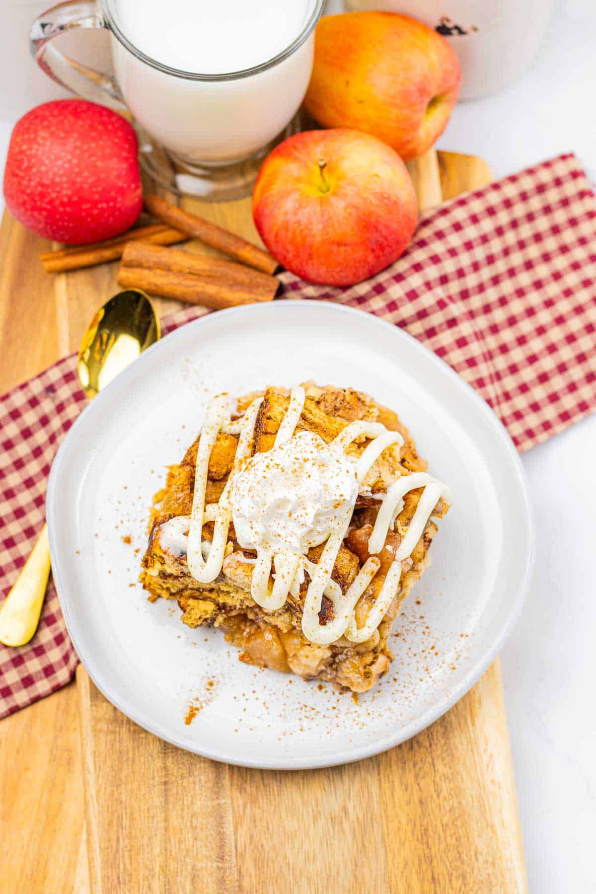 A plate of slow cooker apple pie cinnamon roll casserole with icing and whipped cream, next to apples, cinnamon sticks, and a glass of milk.