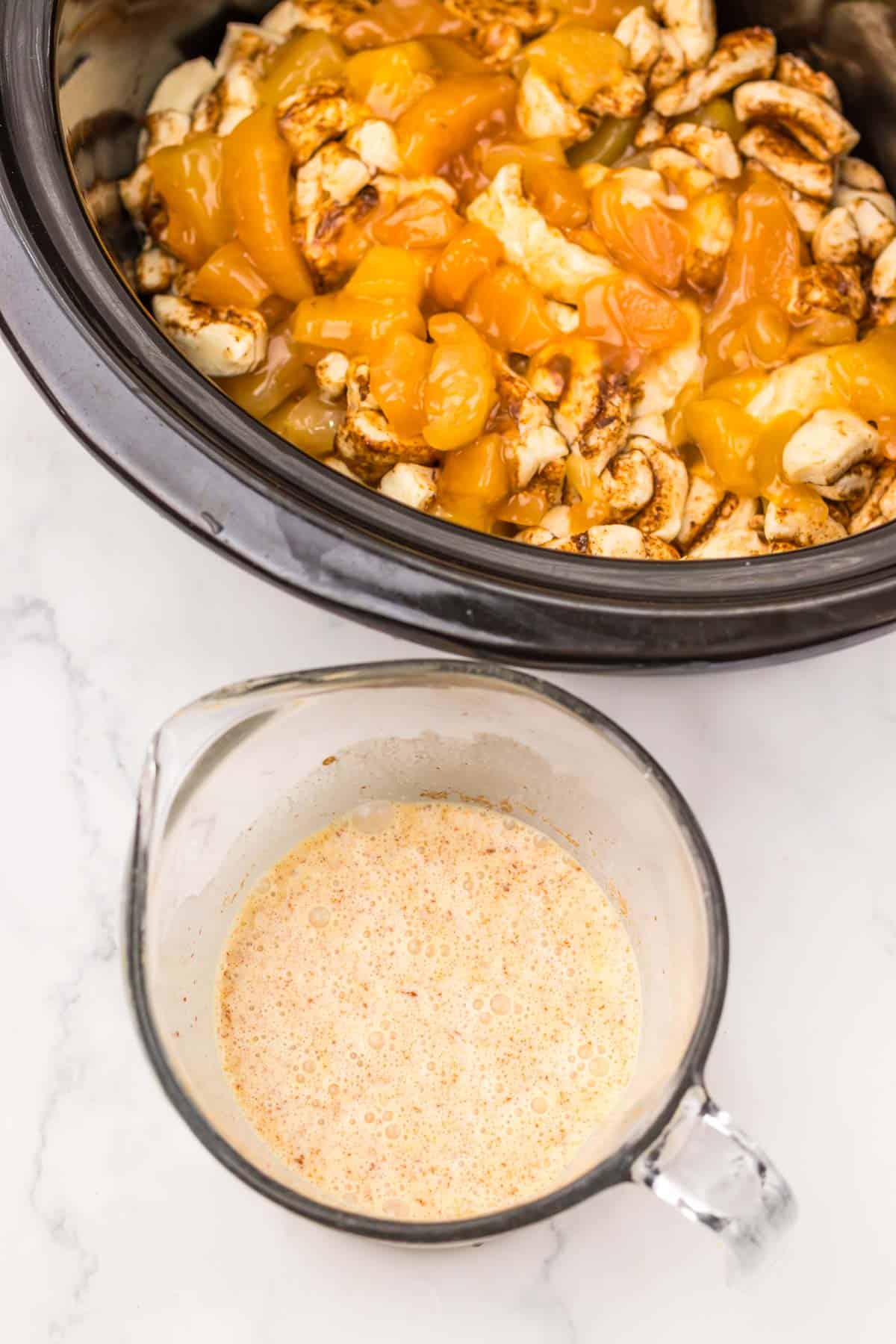 Chopped chicken and peaches in a slow cooker, with a measuring cup of seasoned liquid nearby on a marble counter&mdash;perfect alongside a sweet slow cooker apple pie cinnamon roll casserole.