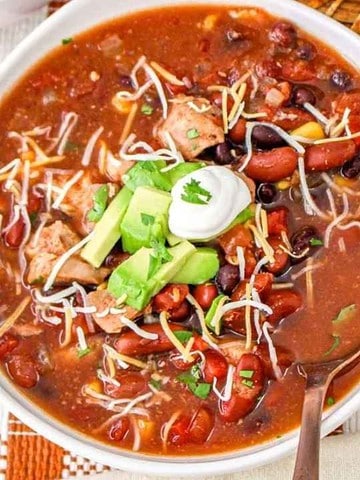 A bowl of chili topped with shredded cheese, avocado, sour cream, and herbs, with lime slices on the side.