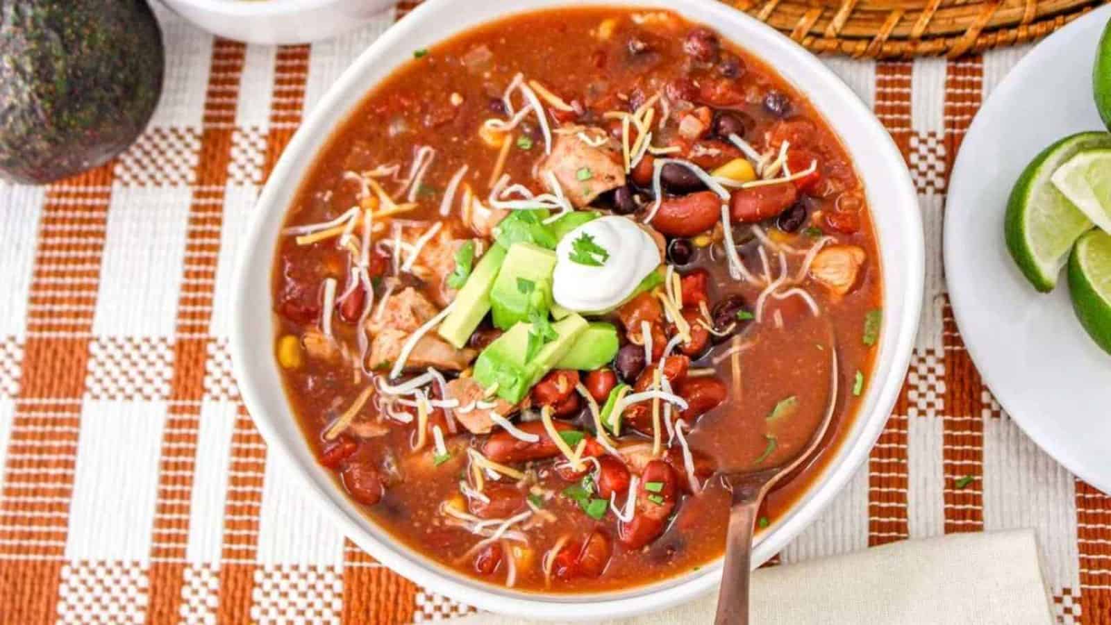 A bowl of chili topped with shredded cheese, sliced avocado, sour cream, and herbs sits on a checkered placemat next to a plate with lime wedges.