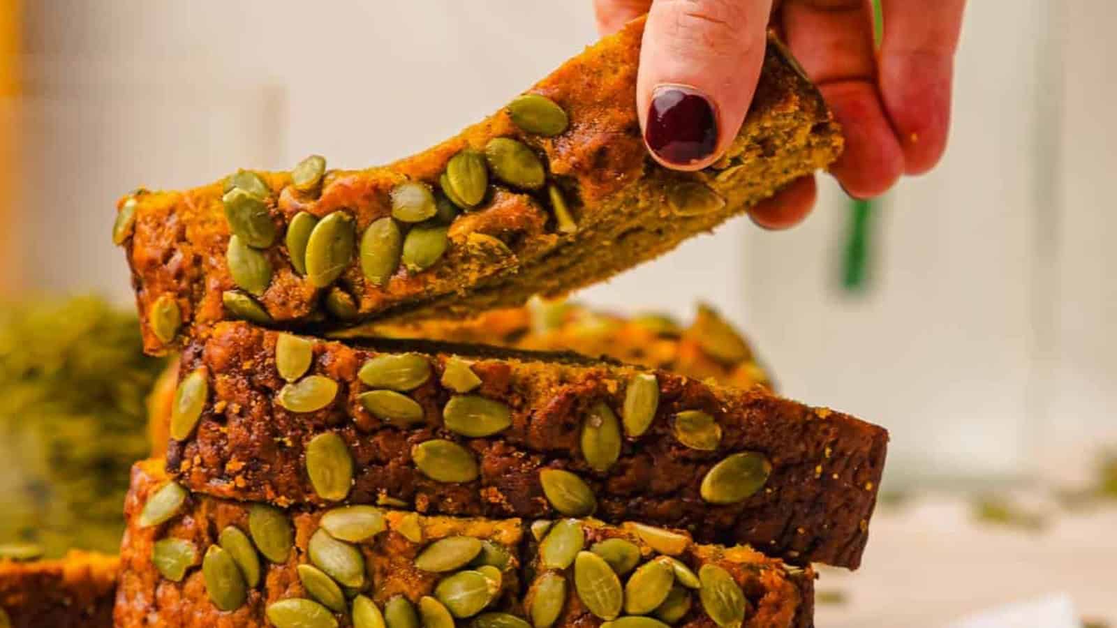 A hand picks up a slice of bread topped with pumpkin seeds from a stack of sliced bread.