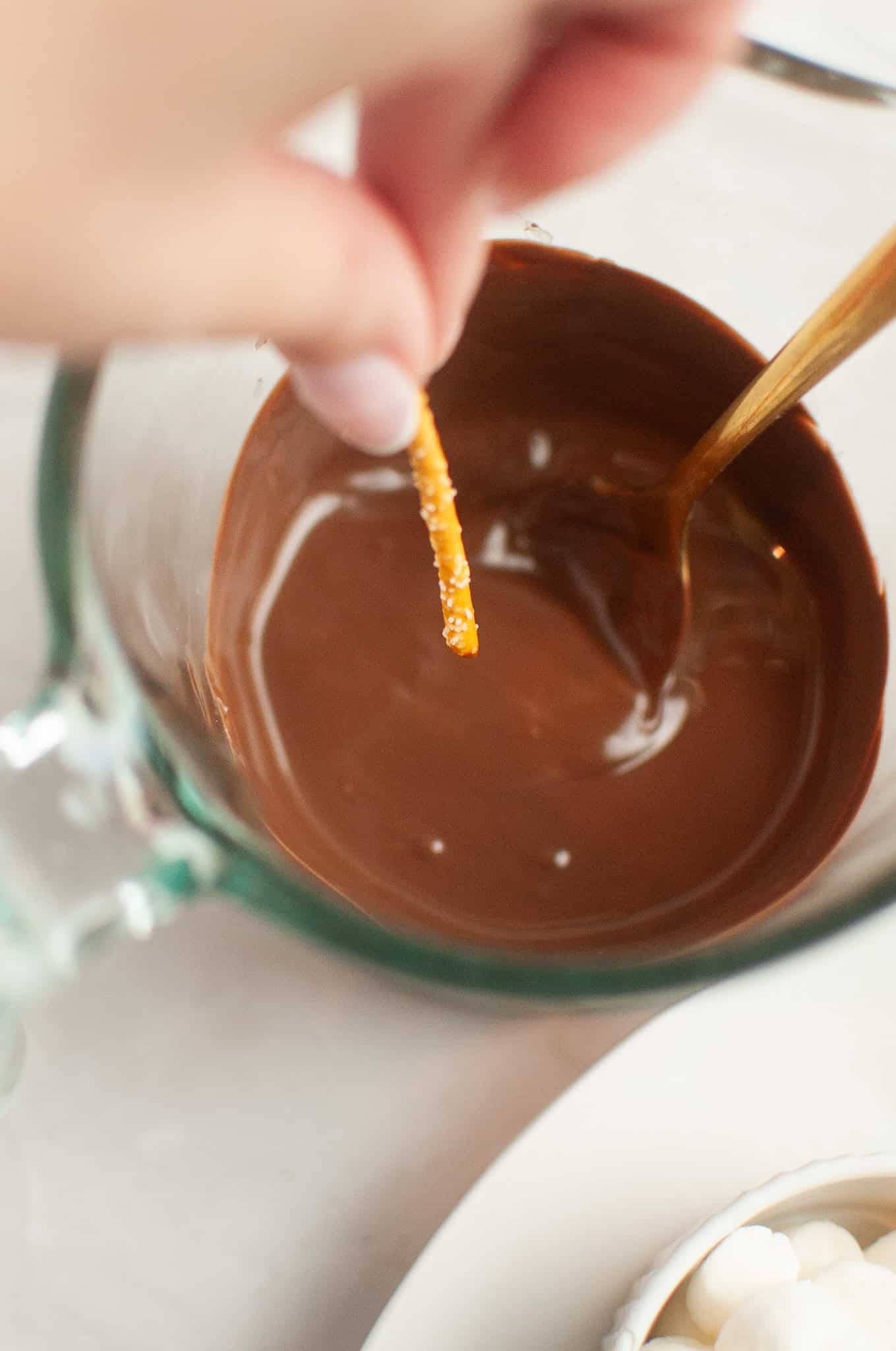 A hand dips a pretzel stick into a bowl of melted chocolate with a spoon beside it, perfect for Thanksgiving treats alongside Turkey and chocolate covered strawberries.