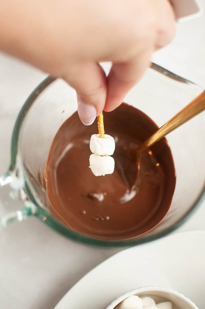 Hand dipping mini marshmallows on a pretzel stick into a bowl of melted chocolate, perfect for creating adorable Thanksgiving turkeys or pairing with chocolate covered strawberry treats.