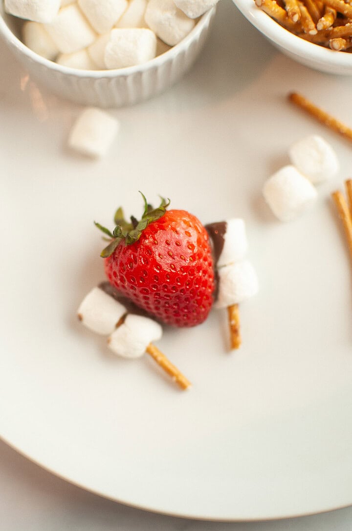 A strawberry surrounded by mini marshmallows and pretzel sticks on a white plate, this playful treat is perfect for Thanksgiving Turkey Desserts or fun takes on Chocolate Covered Strawberries.
