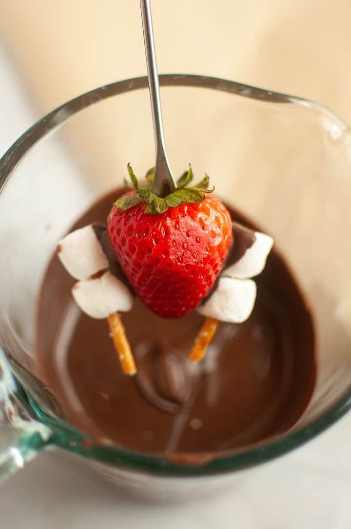 A fork holding a strawberry with marshmallows and pretzels hovers above melted chocolate in a glass cup, perfect for creating Chocolate Covered Strawberries at your next Thanksgiving gathering.