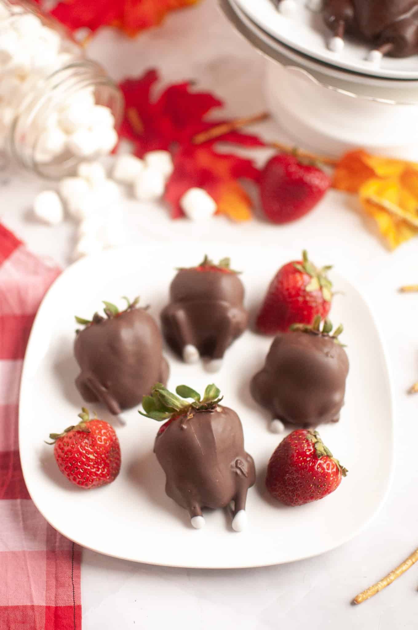 Chocolate Covered Strawberries resembling turkeys with candy feet, perfect for Thanksgiving, are displayed on a white plate alongside plain strawberries.