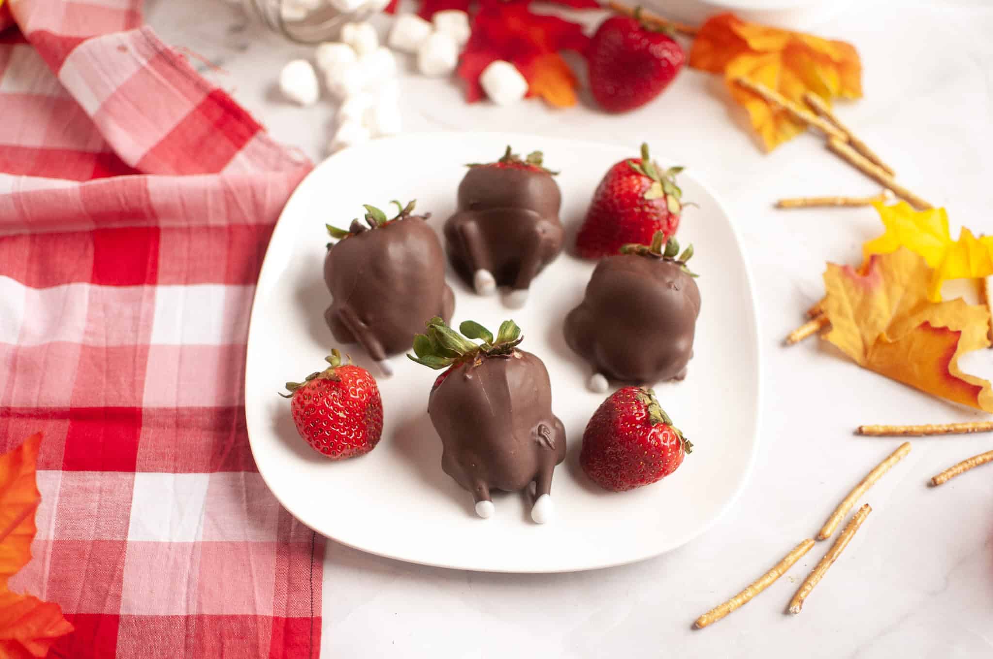 Five Chocolate Covered Strawberries on a plate, some decorated as Turkey Treats, with autumn leaves nearby&mdash;perfect for your Thanksgiving celebration.