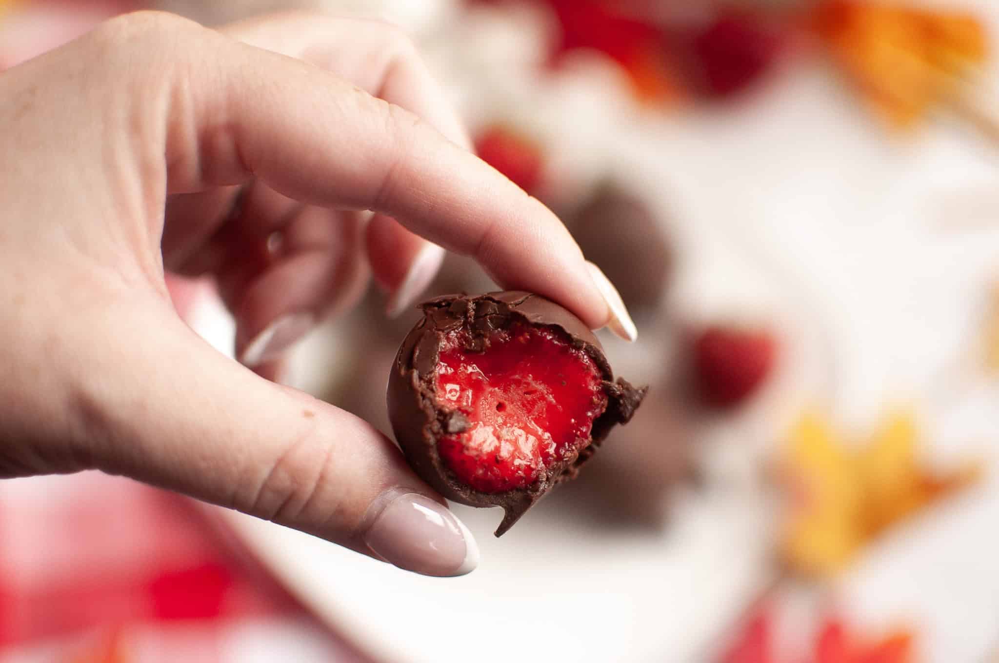 Hand holding a bitten chocolate truffle with a bright red strawberry filling, perfect for Thanksgiving or as a twist on classic Chocolate Covered Strawberries, with a blurred background.