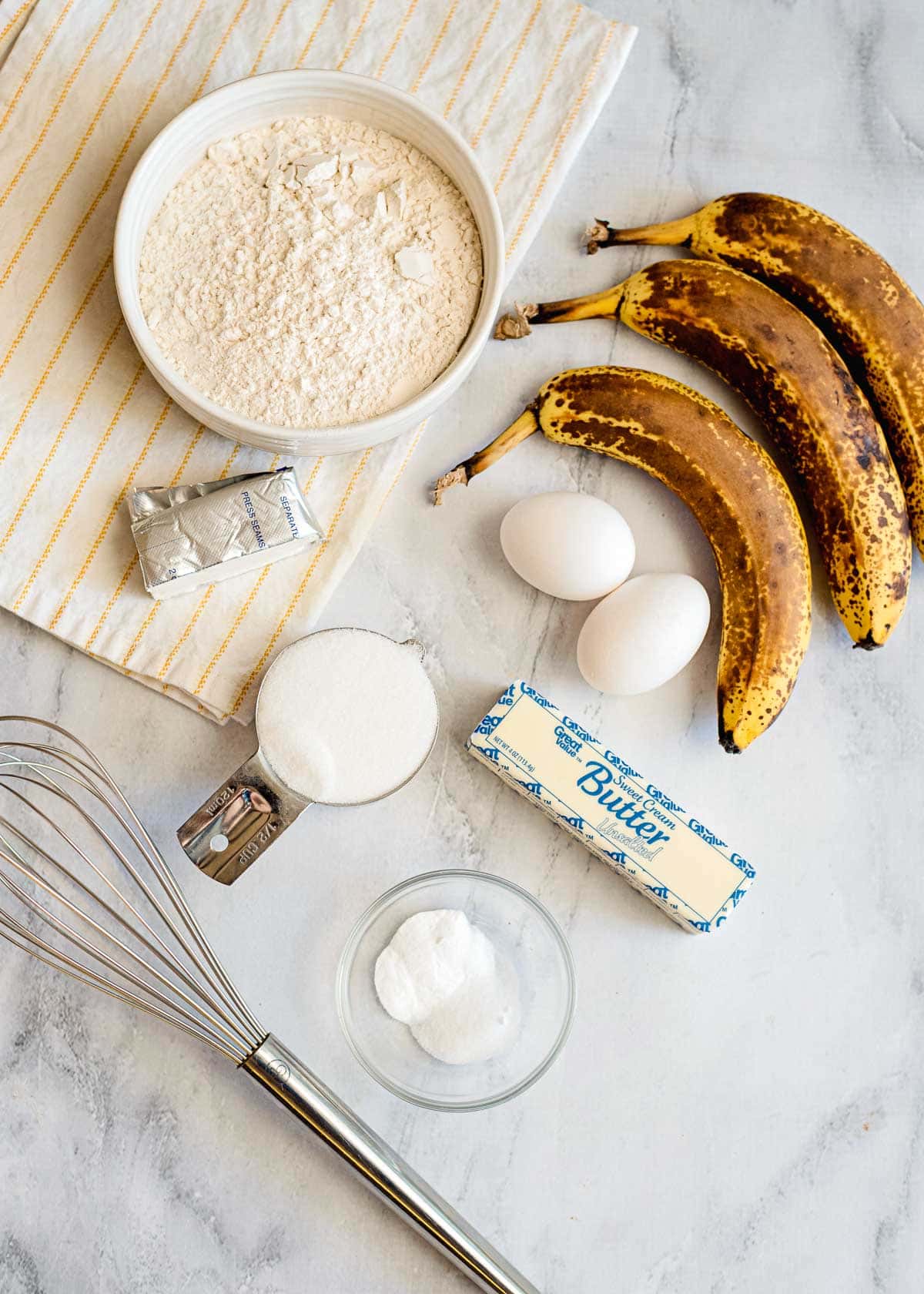 Baking ingredients for banana bread: ripe bananas, flour, eggs, sugar, butter, baking soda, vanilla, and a whisk on a marble surface—perfect for creating a delicious loaf with streusel topping.