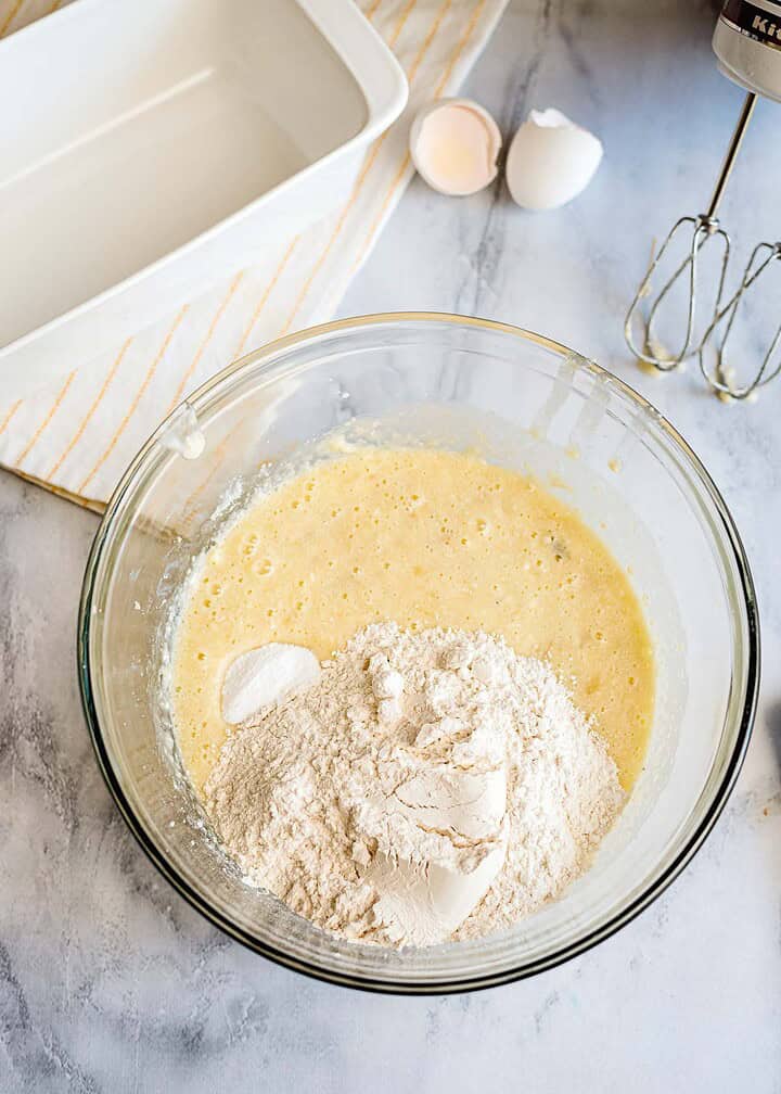 A glass bowl with flour and batter mix for banana bread sits beside a bread pan, cracked eggshells, and a hand mixer on the counter.