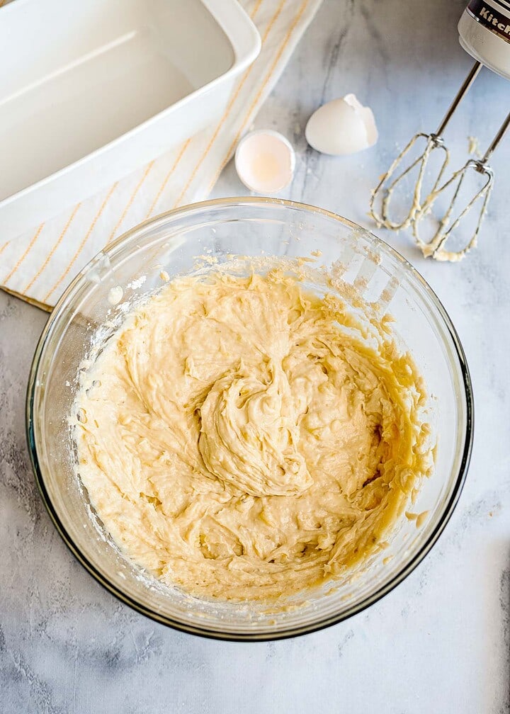 A glass bowl of cake batter sits on a counter beside a cracked egg, a loaf pan, and a hand mixer—perfect for making banana bread or topping with streusel for extra sweetness.