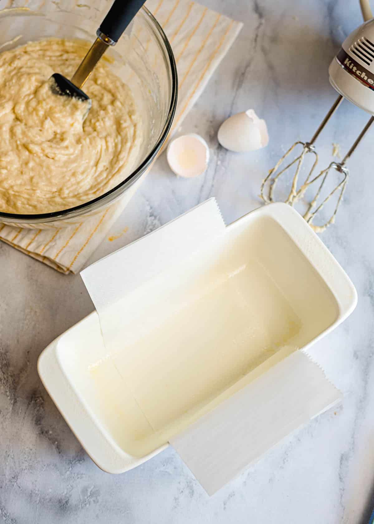 Greased loaf pan lined with parchment paper, next to a bowl of banana bread batter, eggshells, and a mixer—perfect prep for a homemade loaf with optional streusel topping.