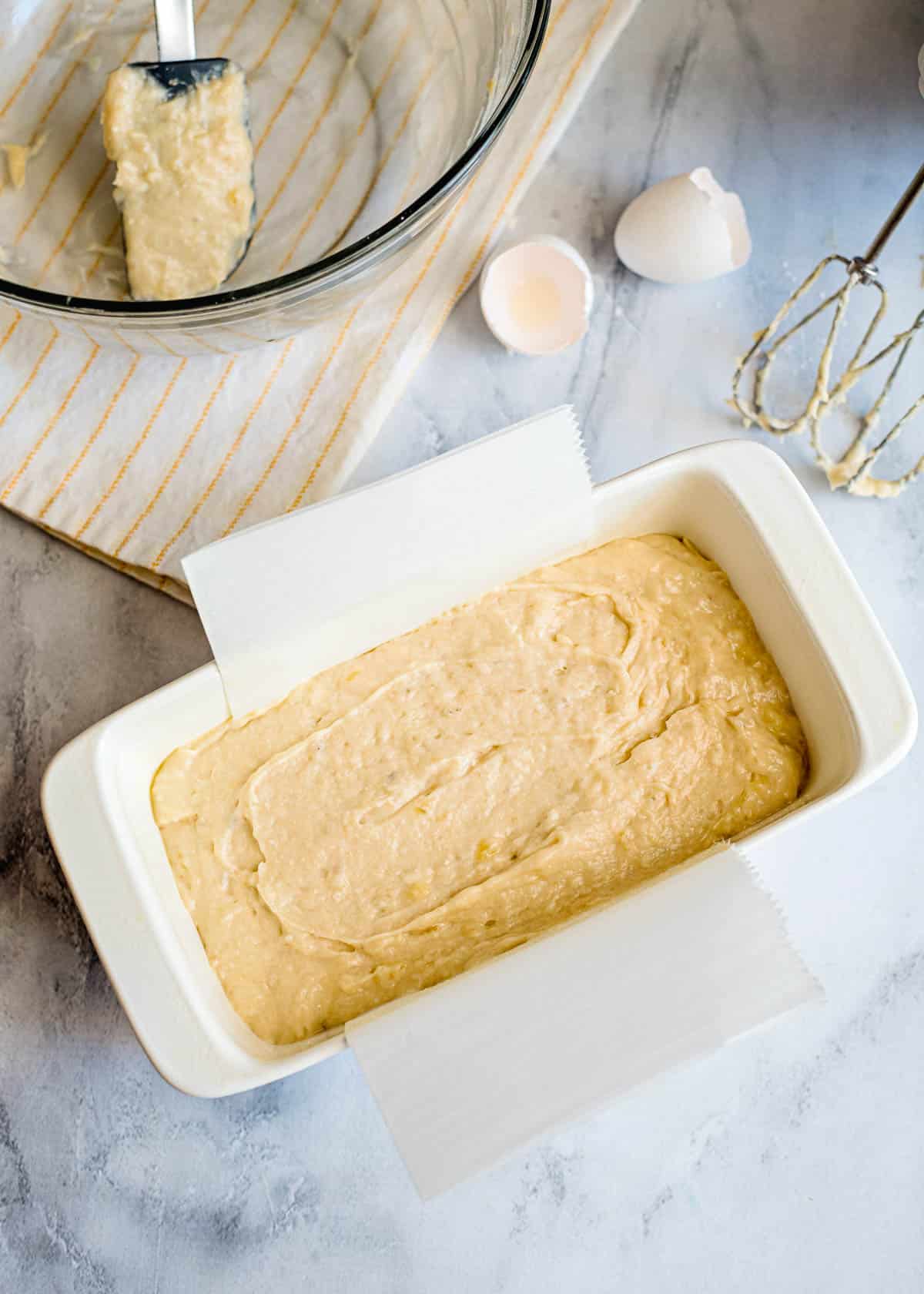 A loaf pan filled with banana bread batter, ready for baking, sits beside mixing tools, eggshells, and a glass bowl—perfect for your favorite banana bread recipe.