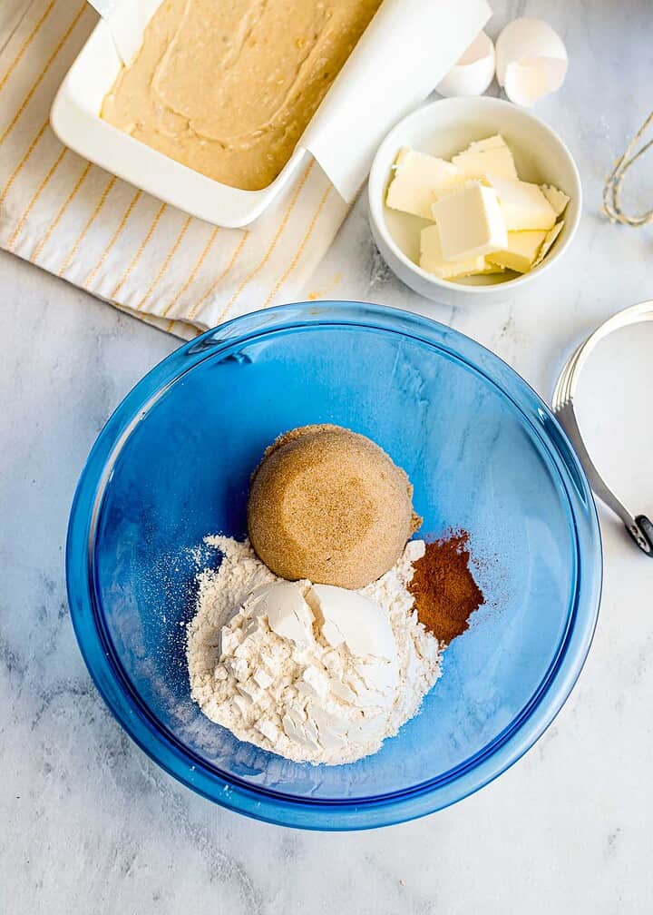 A blue bowl with flour, brown sugar, and cinnamon for a delicious banana bread, alongside butter, eggs, and batter on the kitchen counter—perfect for adding a streusel topping.