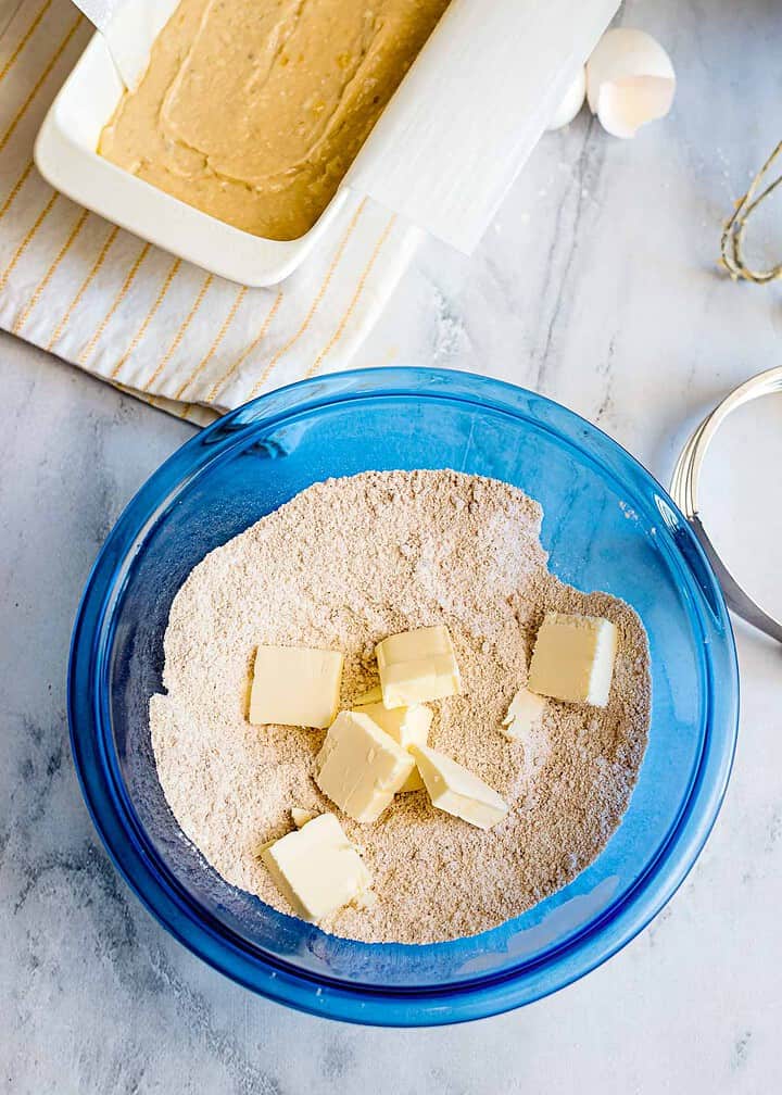 A blue bowl with flour and butter cubes for streusel topping, a baking dish with banana bread batter, and eggs on a marble surface.