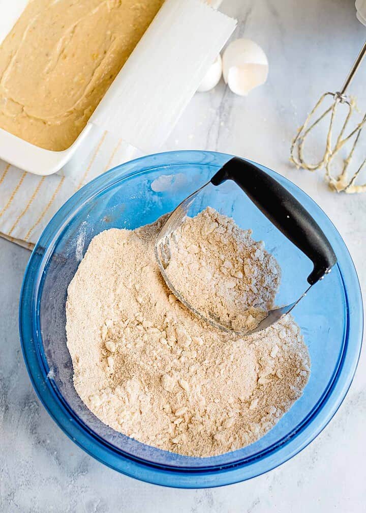 A blue bowl with flour mixture and a pastry cutter, next to cake batter for banana bread, eggshells, and a mixer on a countertop—perfect setup for adding a delicious streusel topping.