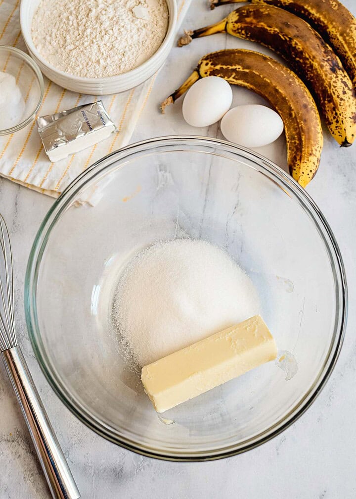 A glass bowl with butter and sugar, surrounded by eggs, overripe bananas, flour, and a whisk on a countertop—perfect for making homemade Banana Bread.