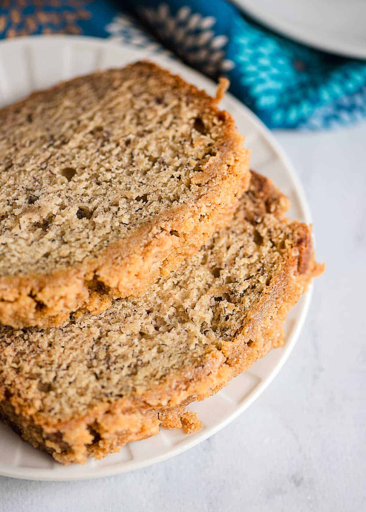 Two slices of banana bread with streusel topping rest on a white plate, set against a blue-patterned cloth in the background.