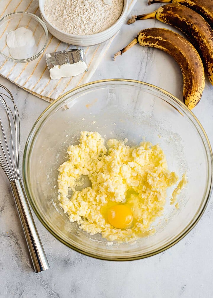 A glass bowl with butter and an egg, whisk, flour, and ripe bananas on a counter—perfect ingredients for homemade banana bread.
